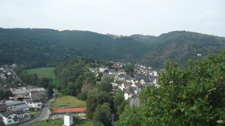 Eine malerische Landschaft mit sanften Hügeln und einem kleinen Dorf. Im Vordergrund sind grüne Felder und Bäume zu sehen.