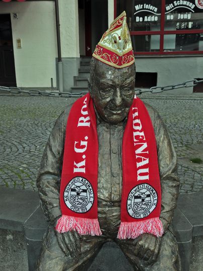 Eine Bronzestatue eines sitzenden Mannes, der einen roten Schal und eine festliche Mütze trägt. Die Statue ist auf einem Pflastersteinplatz in der Stadt aufgestellt.