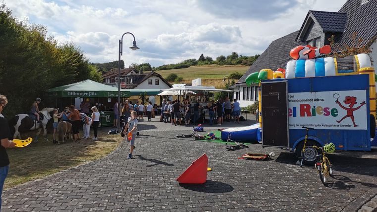 Ein fröhliches Fest mit vielen Leuten und Ständen im Freien. Im Hintergrund sieht man einen bunten Spielwagen und grüne Hügel.