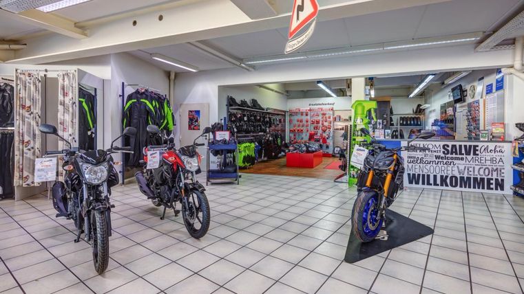 Ein Motorradgeschäft mit mehreren Motorrädern und Motorradbekleidung. Die Wände sind mit verschiedenen Zubehörteilen und Angeboten dekoriert.