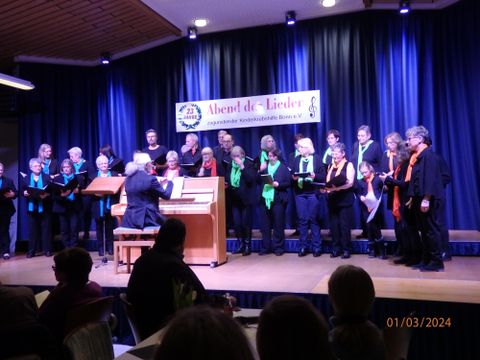 Ein Chor tritt auf einer Bühne auf, während ein Pianist begleitet. Die Sänger tragen verschiedene Farbige Schals und präsentieren ihre Darbietung vor einem Publikum.