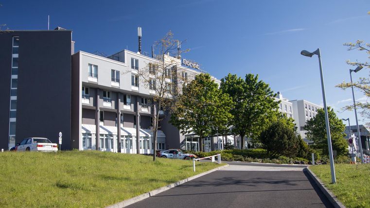 A modern hotel building with clean lines, surrounded by green landscape. The sky is blue and sunny, creating an inviting atmosphere.