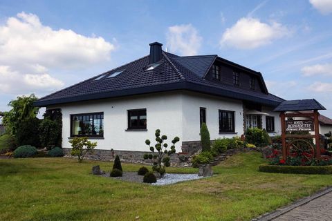 A modern, white house with a black roof and large windows. The well-kept garden is decorated with various plants and a wooden sign.