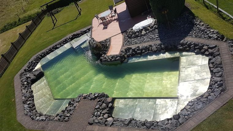 Een modern zwembad met een unieke, gebogen vorm, omringd door stenen en gras. Naast het zwembad staat een gezellige zithoek met een kleine tafel en stoelen.