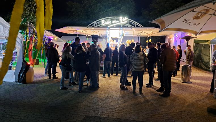 Eine belebte Nachtveranstaltung mit vielen Menschen. Im Hintergrund sieht man ein beleuchtetes Restaurant und Zelte.