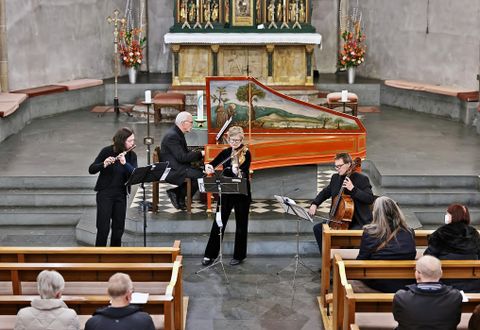 Ein Musikensemble spielt in einer Kirche. Die Zuhörer sitzen auf den Bänken und genießen die Aufführung.