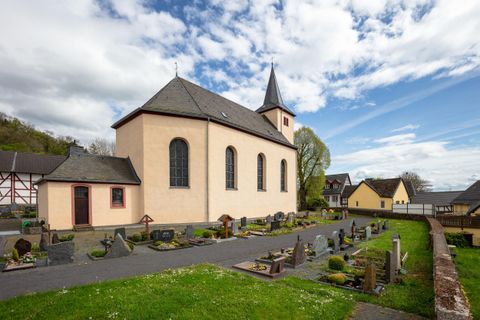Eine Kirche mit einem spitzen Turm und umliegendem Friedhof. Im Hintergrund sind einige traditionelle Häuser zu sehen.