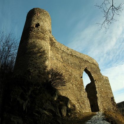 Ruine der Burg Neublankenheim mit rundem Turm und Torbogen, umgeben von kahlen Bäumen, unter blauem Himmel.