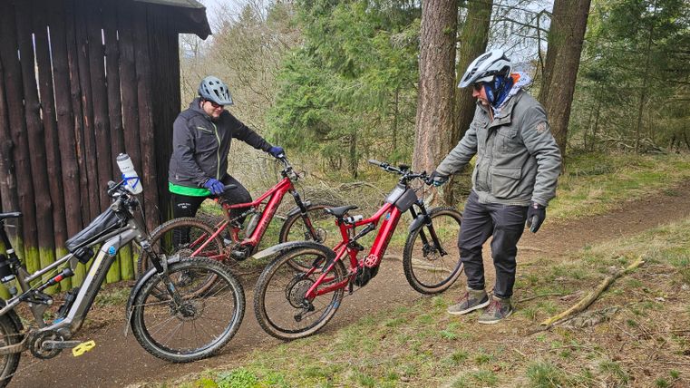Zwei Personen stehen neben ihren E-Bikes auf einem Waldweg. Im Hintergrund sind Bäume und ein Holzschuppen zu sehen.