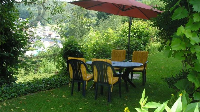 Een gezellige tuintafel met stoelen en een parasol staat in een groene omgeving. Op de achtergrond zijn bomen en een klein dorp zichtbaar.
