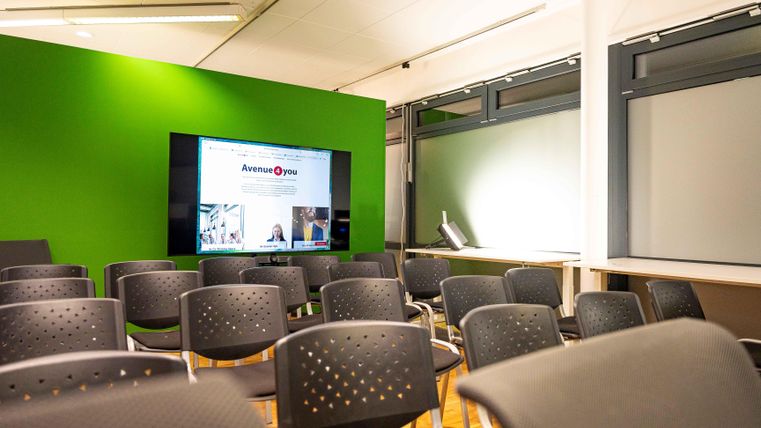 Ein moderner Raum mit einer grünen Wand und einer großen Bildschirmpräsentation. Vorne stehen Reihen von schwarzen Stühlen, die auf eine Veranstaltung warten.