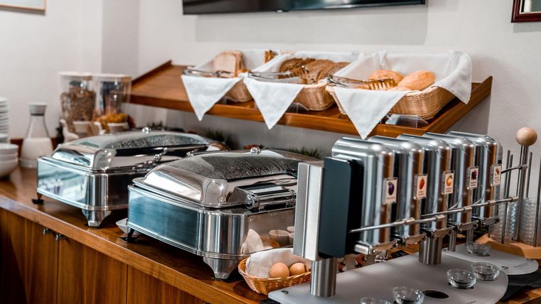 Een ontbijtbuffet met verschillende warme gerechten in grote containers. In de bovenste rij zijn verse broodjes en gebak gerangschikt.