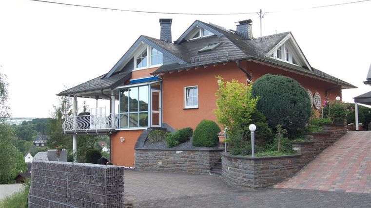A beautiful orange house with a balcony and a well-maintained garden. The driveway is decorated with stones, green plants, and trees.