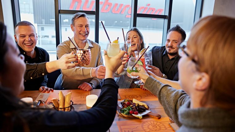 A group of friends is sitting at a table and raising their glasses for a toast. In the background, large windows with a striking inscription can be seen.