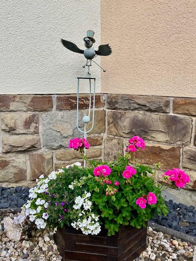 Une jardinière colorée avec des fleurs roses et blanches. Au-dessus se trouve une statue décorative d'oiseau avec un chapeau.