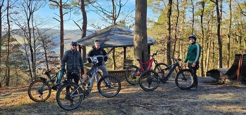Gruppe von Radfahrern in einem Waldgebiet mit Fahrrädern. Im Hintergrund ist eine kleine Hütte und Bäume zu sehen.