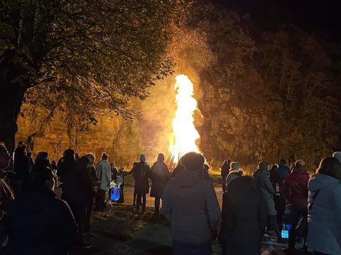 Eine Gruppe von Menschen steht um ein großes Feuer herum. Die Nacht ist dunkel und die Flamme erhellt die Umgebung.