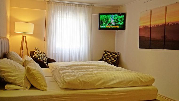 Une chambre confortable avec un grand lit et un système de télévision moderne. Les fenêtres sont décorées de rideaux clairs et il y a un fauteuil dans un coin.