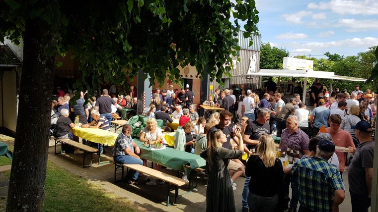 Eine lebhafte Veranstaltung im Freien mit vielen Menschen, die an Tischen sitzen und sich unterhalten. Die Atmosphäre ist freundlich und gesellig, umgeben von Bäumen und Sonnenschein.