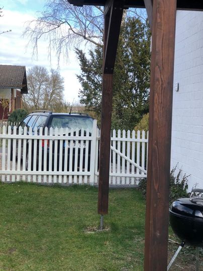 A car is parked behind a white picket fence in a well-kept garden. In the foreground, a gazebo and a grill are visible.