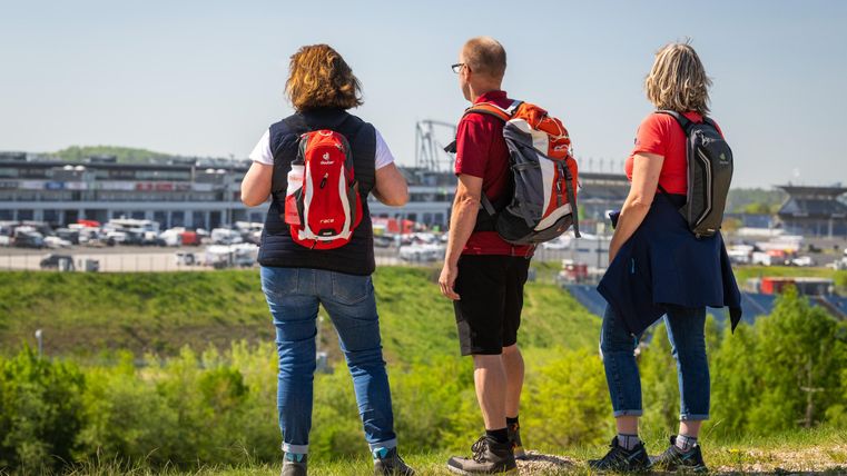 Drei Personen stehen am Hang und schauen auf ein Stadion. Sie tragen Rucksäcke und leichte Kleidung, ideal für einen Ausflug.