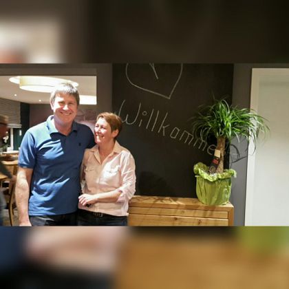 A couple stands in an inviting room in front of a wall with the inscription "Welcome". In the background, a plant and a wooden table are visible.