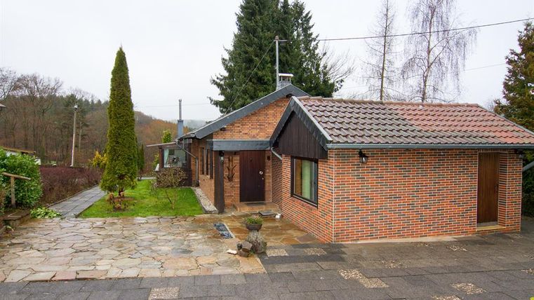 A modern house made of red bricks with a green front yard. In the background, trees and a tranquil environment can be seen.
