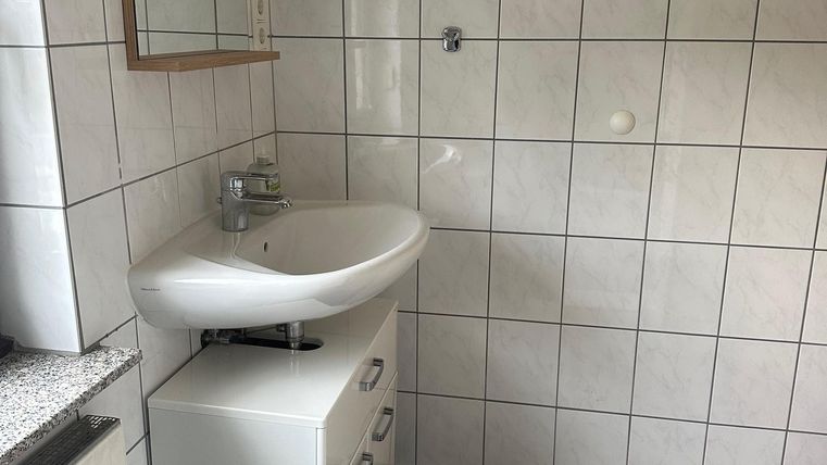 Een moderne badkamer met witte tegels en een wastafel op een klein onderkastje. De ruimte is licht en eenvoudig ingericht, met een spiegel boven de wastafel.