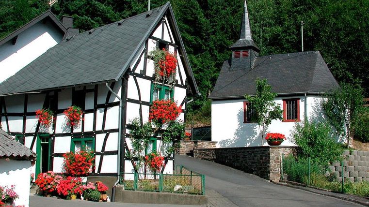 Ein malerisches Dorf mit traditionellen Fachwerkhäusern und einer kleinen Kirche. Bunte Blumen schmücken die Gebäude und die Umgebung ist grün und einladend.