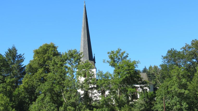 Eine Kirche mit einem spitzen Turm ist zwischen Bäumen sichtbar. Der Himmel ist klar und blau.