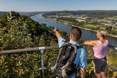 Zwei Personen stehen auf einer Aussichtsplattform über dem Rheinflusstal. Sie schauen auf den breiten Fluss, die Stadt am Ufer und die bewaldeten Hügel. Eine Person zeigt mit dem Arm in die Landschaft. Vor ihnen liegen grüne Hänge, die sich bis zum Wasser hinunterziehen.