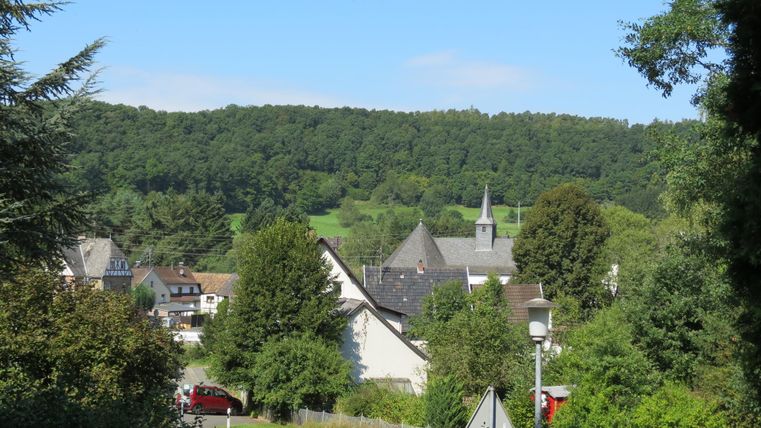 Ein malerisches Dorf umgeben von üppigem Grün und sanften Hügeln. Im Hintergrund ist eine Kirche mit einem hohen Turm zu sehen.