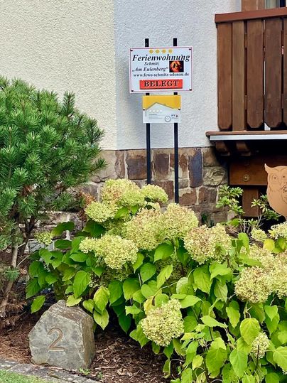 Un panneau indiquant un appartement de vacances avec l'inscription "Appartement de vacances au Entenbach". Au premier plan, on voit des plantes vertes et une pierre avec le numéro "2".