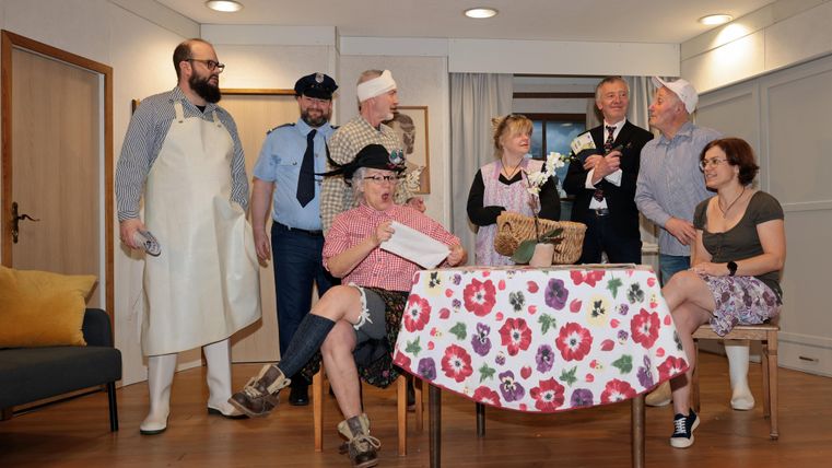 Eine Gruppe von Menschen in verschiedenen Kostümen steht und sitzt um einen Tisch. Sie scheinen eine Szene aus einem Theaterstück oder einem Sketch zu repräsentieren.