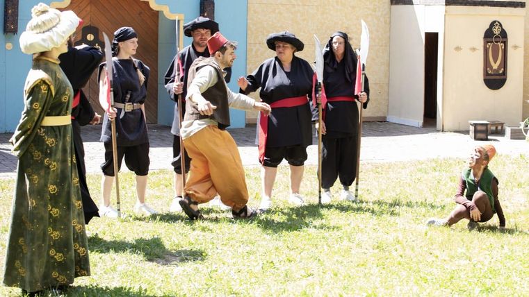 An outdoor scene performance with several actors in historical costumes. In the foreground, there is a person making a dramatic gesture while other spectators watch.