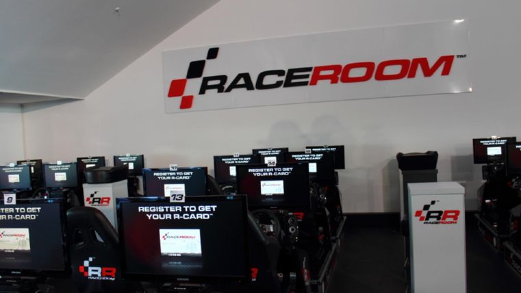 Ein moderner Racing-Simulator-Bereich mit mehreren Spielstationen. Im Hintergrund ist das Logo von RaceRoom zu sehen.
