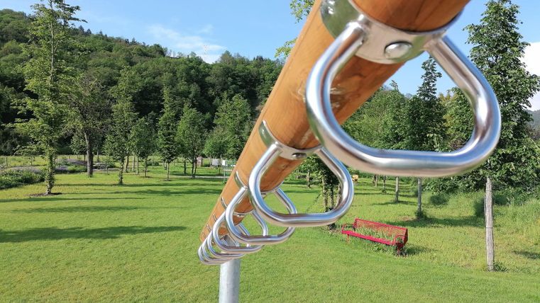Ein Holzgestell mit Metallringen steht in einem grünen Park. Im Hintergrund sieht man Bäume und eine Bank auf dem Rasen.