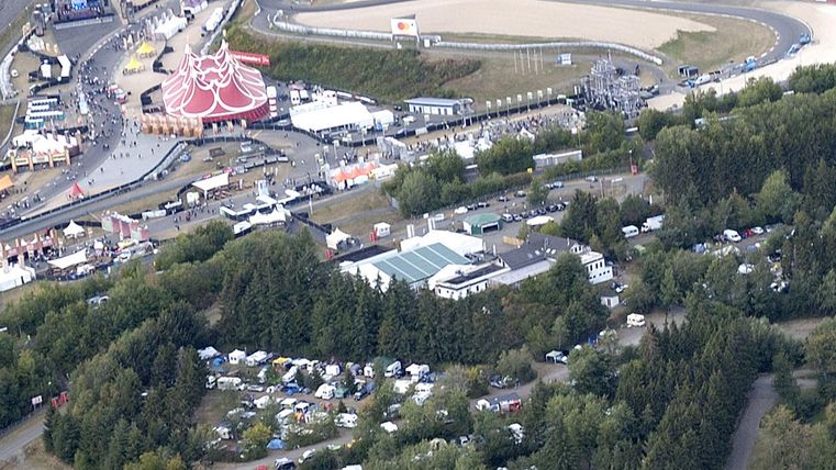 An overview of a racetrack area with various vehicles and tents. In the background, racetracks and an event area can be seen.