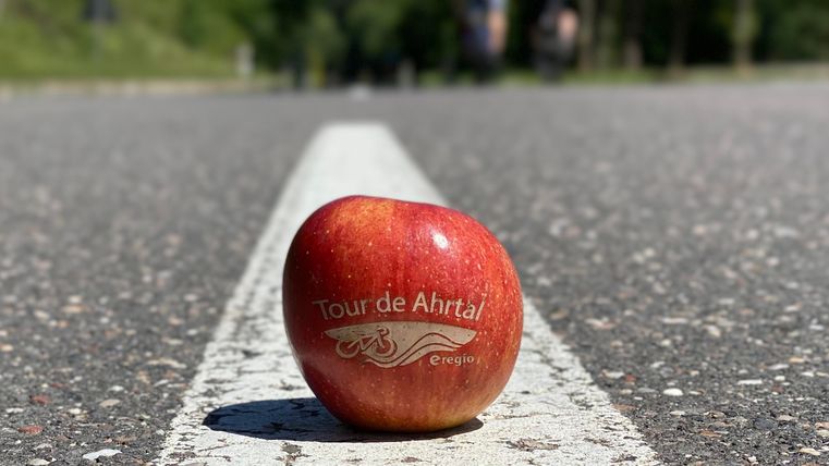 Ein roter Apfel liegt auf der Straße, gekennzeichnet mit "Tour de Ahrtal". Im Hintergrund sind verschwommen Radfahrer zu sehen.