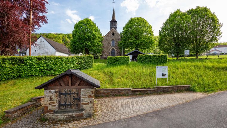 Een charmante kerk omringd door groene bomen en weelderig gras. In de voortuin bevindt zich een kleine stenen sokkel met een houten dak.