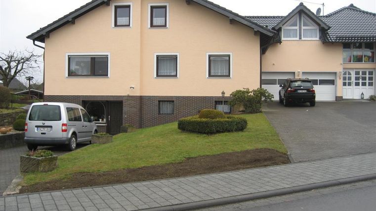 Ein großes, zweigeschossiges Haus mit einer hellen Fassade. Es gibt eine Auffahrt mit zwei Autos und einen gepflegten Vorgarten.