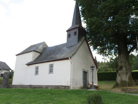 A small church with a pointed tower and a bright entrance. Surrounded by green spaces and trees.