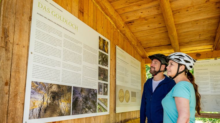 Zwei Radfahrer stehen unter einem Holzüberstand und betrachten Informationstafeln. Sie tragen Helme und lächeln, während sie die Texte lesen.