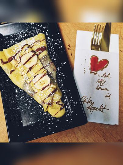 Une délicieuse crêpe avec des bananes, de la sauce au chocolat et du sucre glace sur une assiette noire. À côté se trouve une serviette avec un message chaleureux.