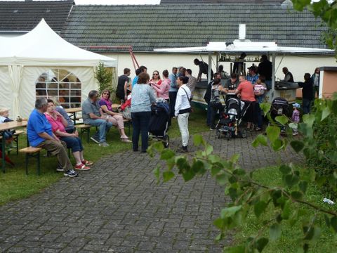 Eine gesellige Veranstaltung im Freien mit vielen Menschen und Kinderwagen. Im Hintergrund ist ein Verkaufsstand und ein Zelt zu sehen.