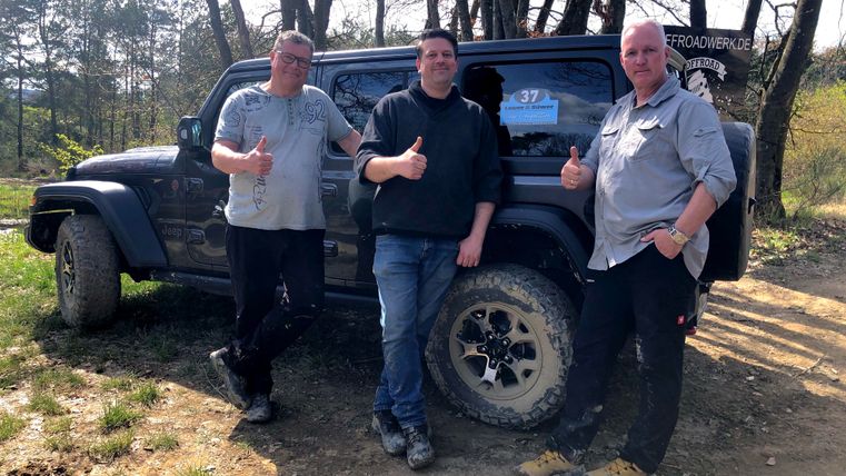 Drei Männer stehen lächelnd neben einem geländefähigen Fahrzeug im Freien. Sie geben Daumen hoch und die Umgebung ist grün und baumreich.