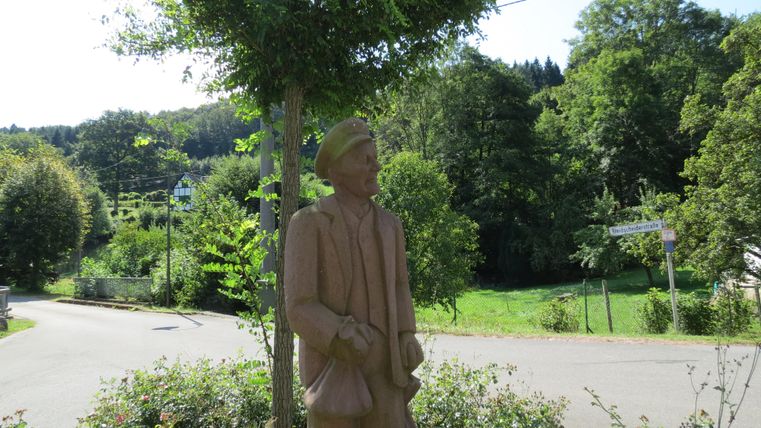 Eine Statue eines Mannes steht an einer Straßenecke, umgeben von Bäumen und Grün. Im Hintergrund sind sanfte Hügel und ein klarer Himmel zu sehen.