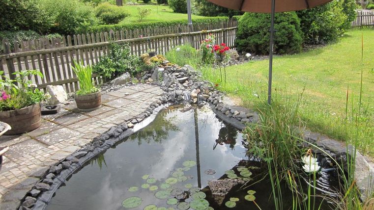 Een kleine vijver met waterlelies en planten omringd door een verzorgde tuin. Een parasol biedt schaduw op het terras.