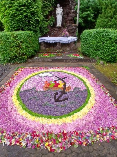 Een kunstzinnig bloemenpatroon op de vloer, omringd door groene struiken. In het midden bevindt zich een standbeeld en kleurrijke bloesems in de vorm van landschappen.