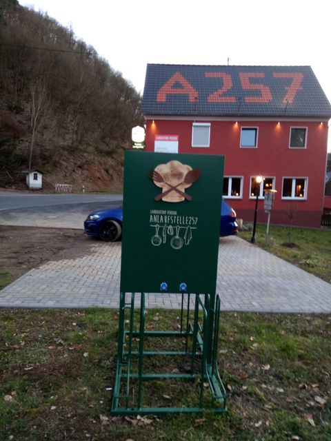 A green box with a blooming motif is standing in a parking lot. In the background, there is a red building with the inscription "A257".